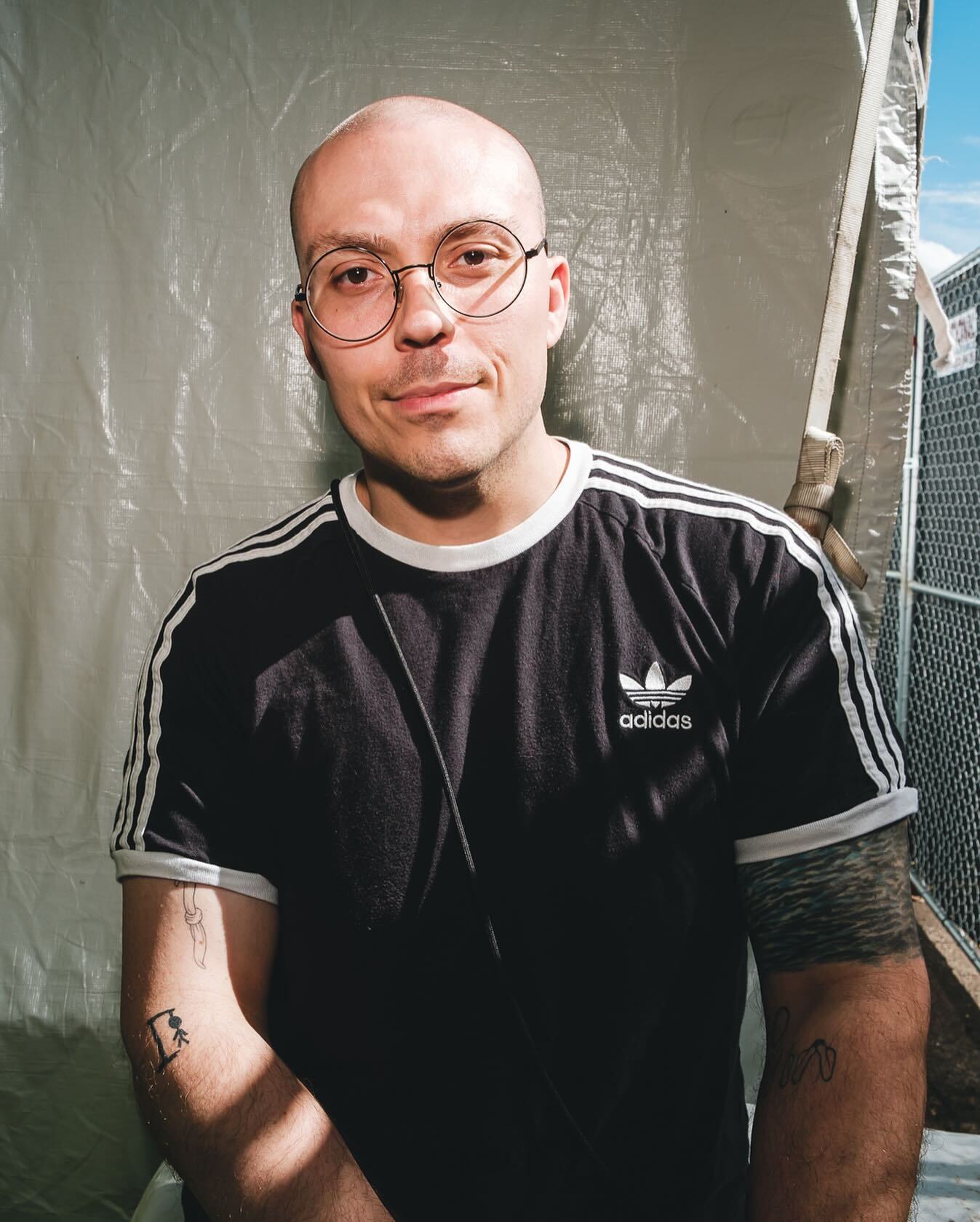 Anthony Fantano, bald with round wire-frame glasses, wearing a black Adidas shirt, photographed outdoors.